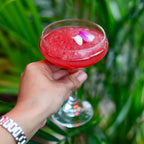 Hand holding a red cocktail with a flower against a green leafy background
