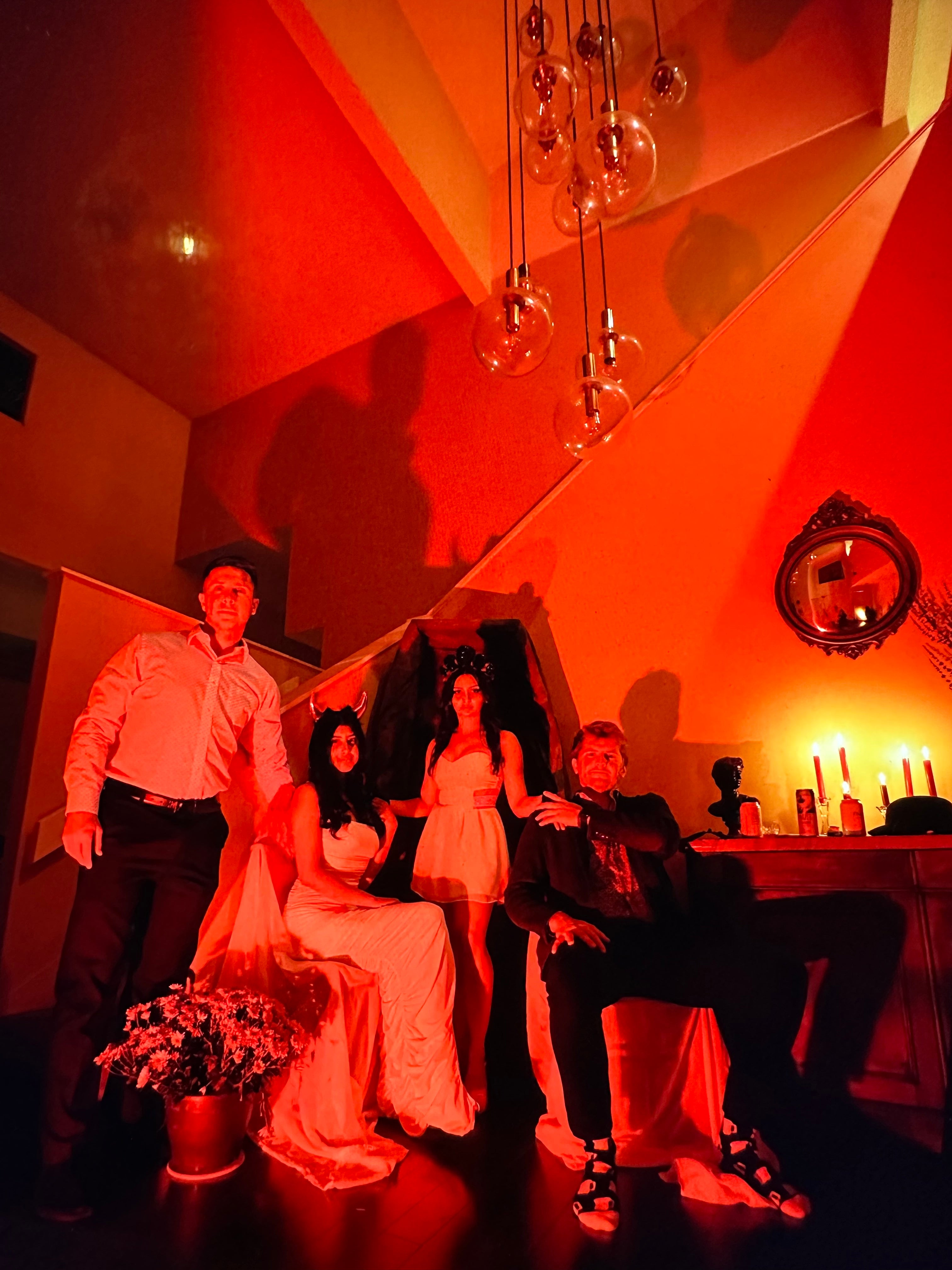 Four people posing together in a dimly lit attic or room with dramatic red and orange lighting. Two women are in wedding-style gowns, one standing and one seated, while a man in a white shirt and dark pants stands on the left and another man in dark clothing sits on the right.