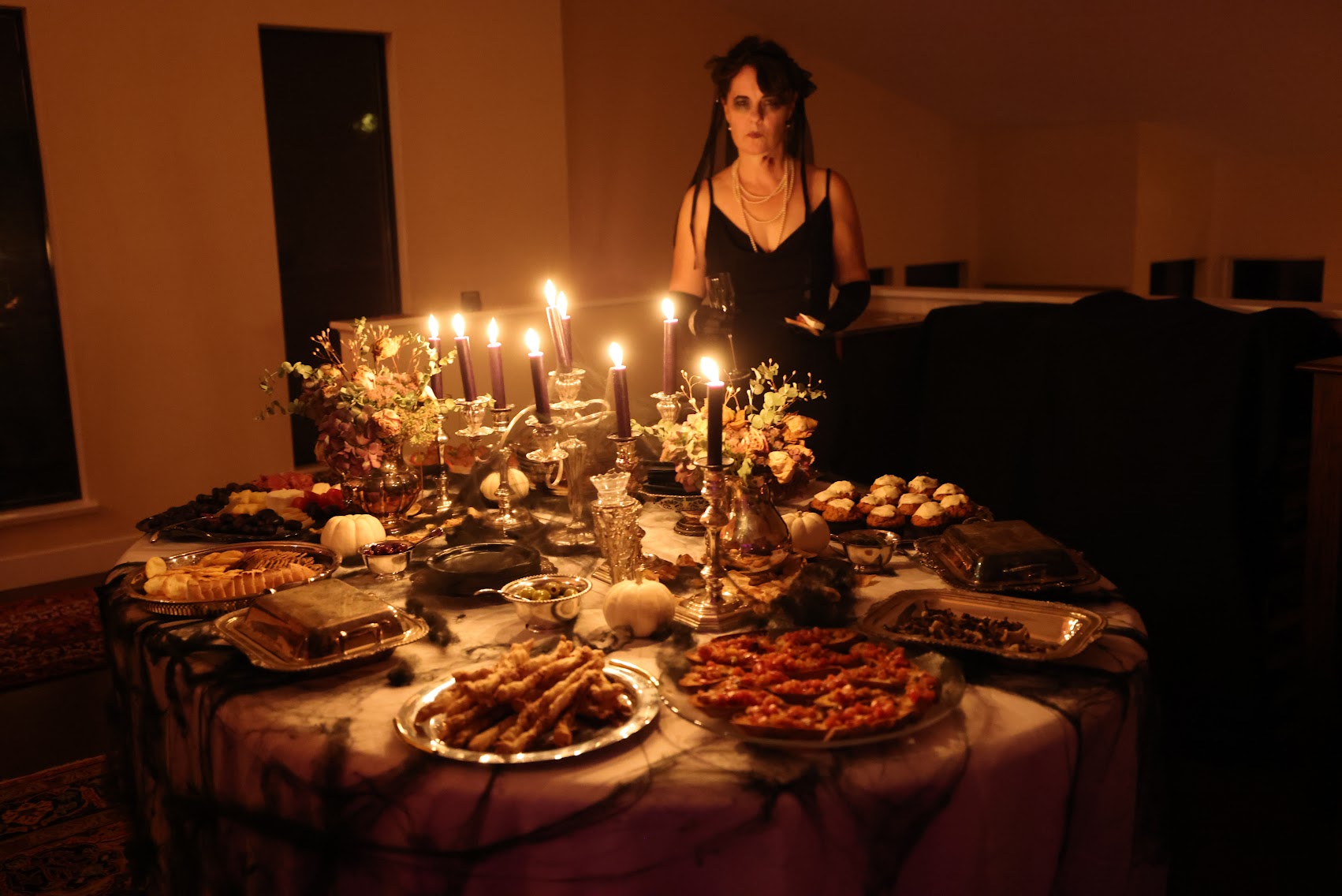 A woman in a black dress stands behind a heavily decorated dining table lit by many candles in a dimly lit room. The table is covered with food, silverware, and floral arrangements.