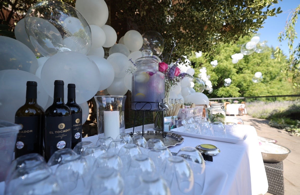 An outdoor catering table decorated with white balloons, wine bottles, a glass beverage dispenser, and clear wine glasses on a white tablecloth, set under trees.