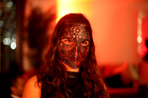 A woman with long wavy brown hair wearing an ornate black lace mask over her face, photographed in warm orange-red ambient lighting with a blurred indoor background.
