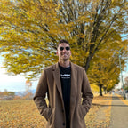 Man wearing a brown coat and sunglasses standing in front of a tree with yellow leaves.