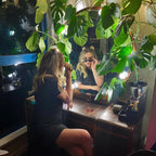 Person sitting at a vanity mirror surrounded by plants, wearing a black dress and sunglasses.