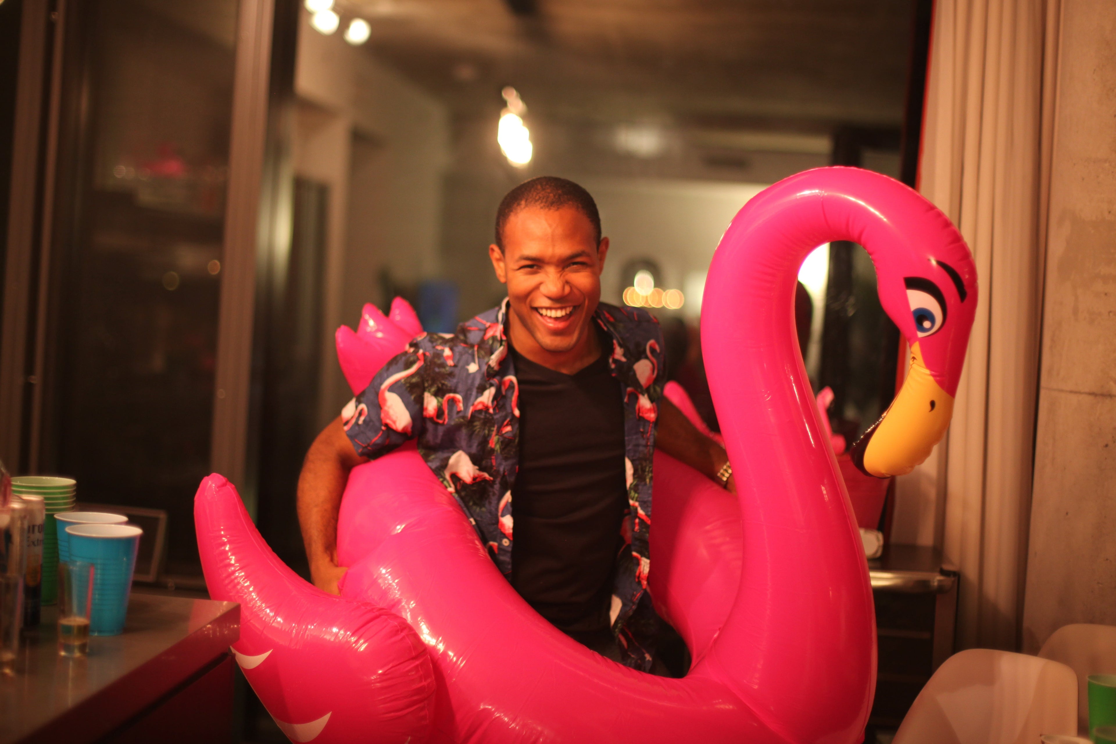 A man smiling while sitting inside a large pink inflatable flamingo float in a dimly lit indoor setting.