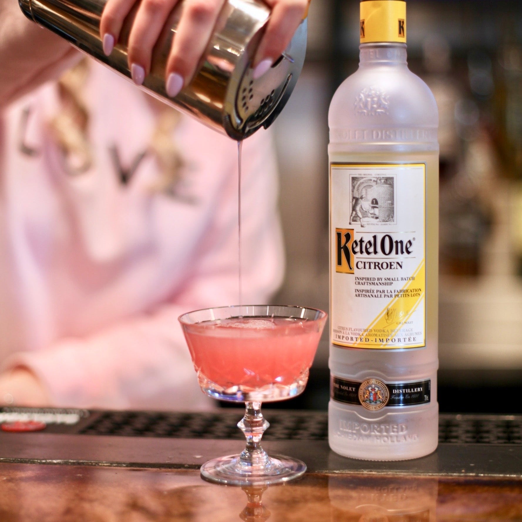 Bottle of Ketel One vodka with a cocktail being poured into a glass on a bar.