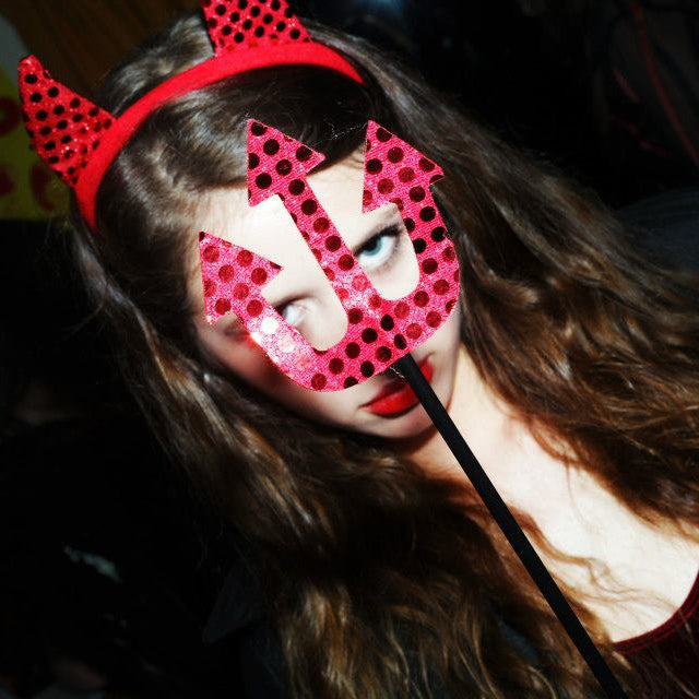 Person wearing a red devil horn headband and a pink eye mask with a black stick.