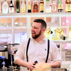 Man in a white shirt and suspenders standing in a bar with shelves of alcohol behind him