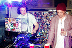 Two men at a DJ booth in a club or event space, with one operating turntables and a laptop covered in stickers, and the other holding a drink. The background is a collage of many small photos under purple and blue lighting.