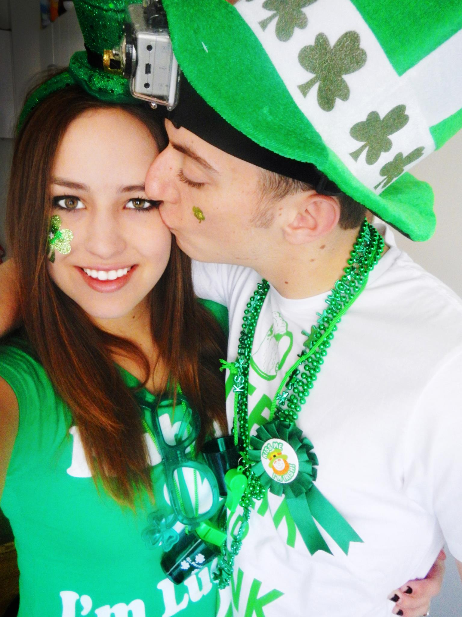 A couple celebrating St. Patrick's Day, with the woman wearing a green shirt and shamrock face paint while the man kisses her on the cheek.