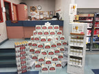 A retail display of Stella Artoisan coffee and tea products arranged in stacks and on a freestanding brick-style shelf inside a store. Visible Stella Artoisan labels appear on many cartons and cans, and the setting includes a counter, bulletin board, and refrigerator in the background.