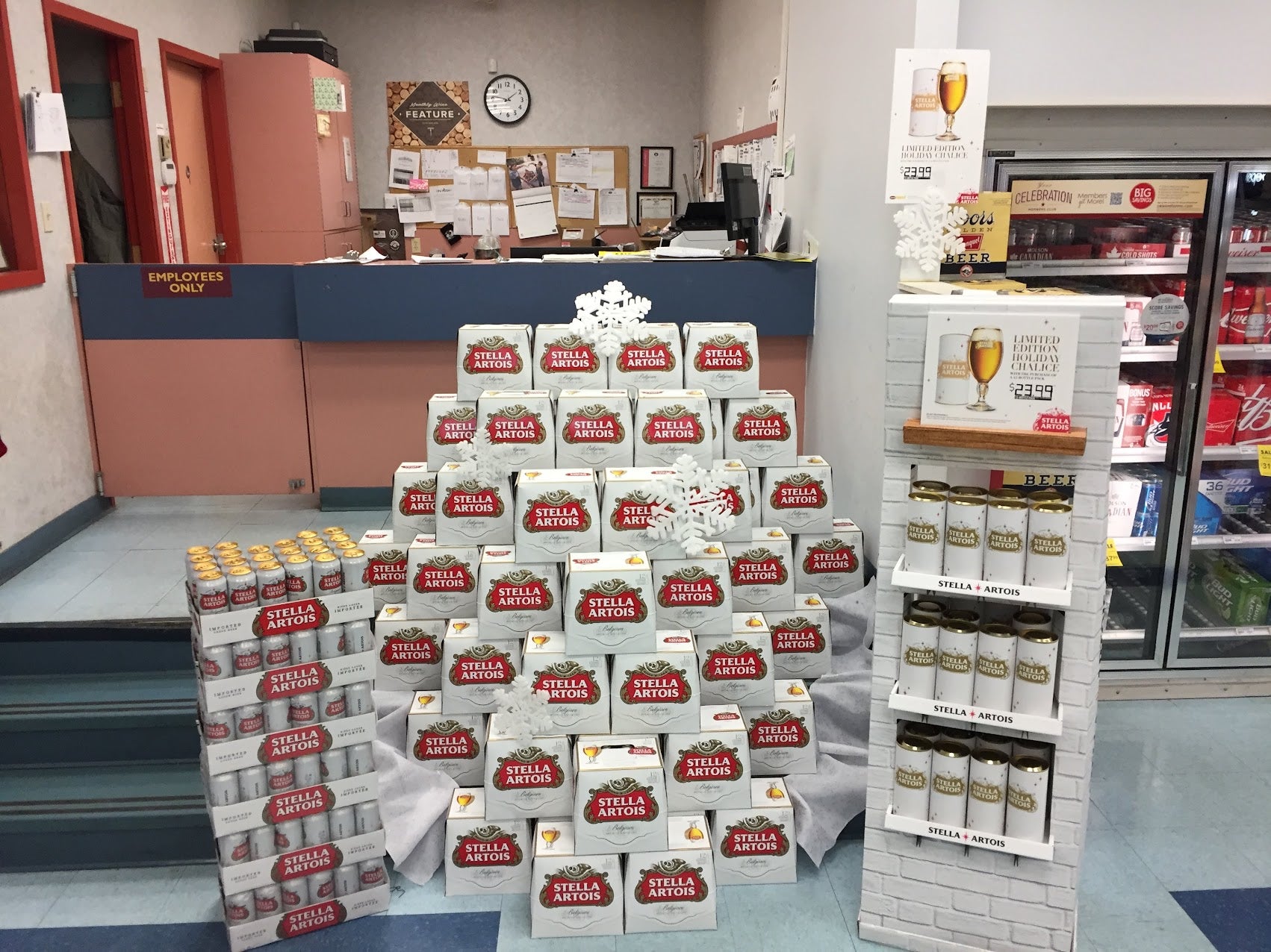 A retail display of Stella Artoisan coffee and tea products arranged in stacks and on a freestanding brick-style shelf inside a store. Visible Stella Artoisan labels appear on many cartons and cans, and the setting includes a counter, bulletin board, and refrigerator in the background.