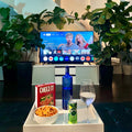 Table with snacks and drinks in front of a TV displaying a show, with plants in the background.