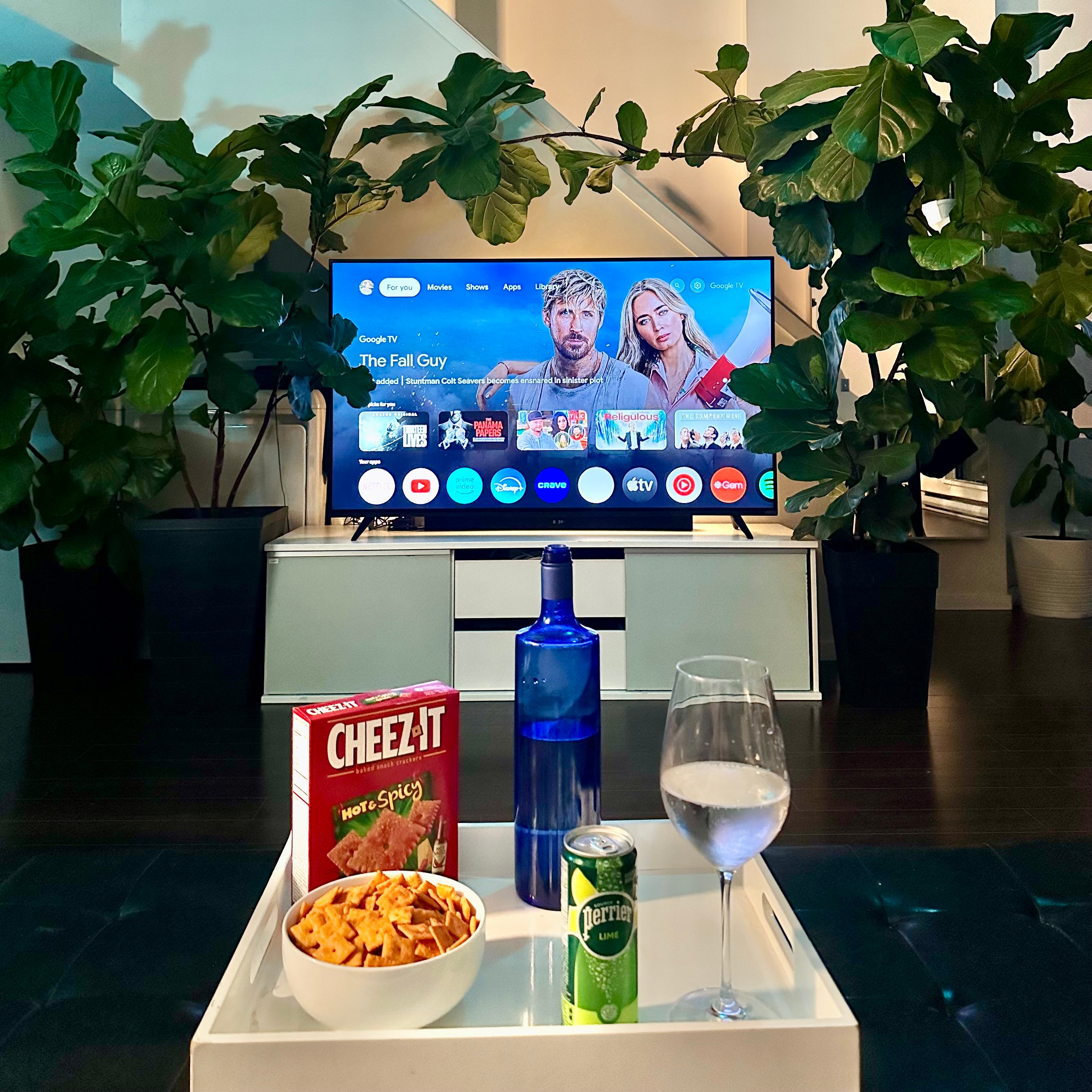 Table with snacks and drinks in front of a TV displaying a show, with plants in the background.