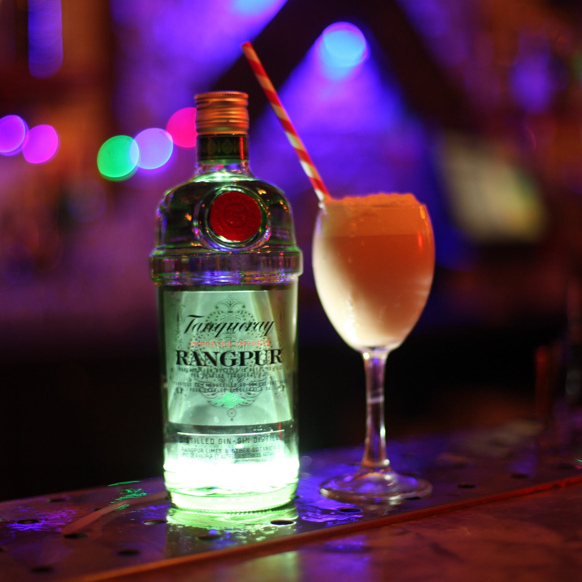 Bottle of Tanqueray Rangpur gin with a cocktail on a bar counter with colorful lights in the background