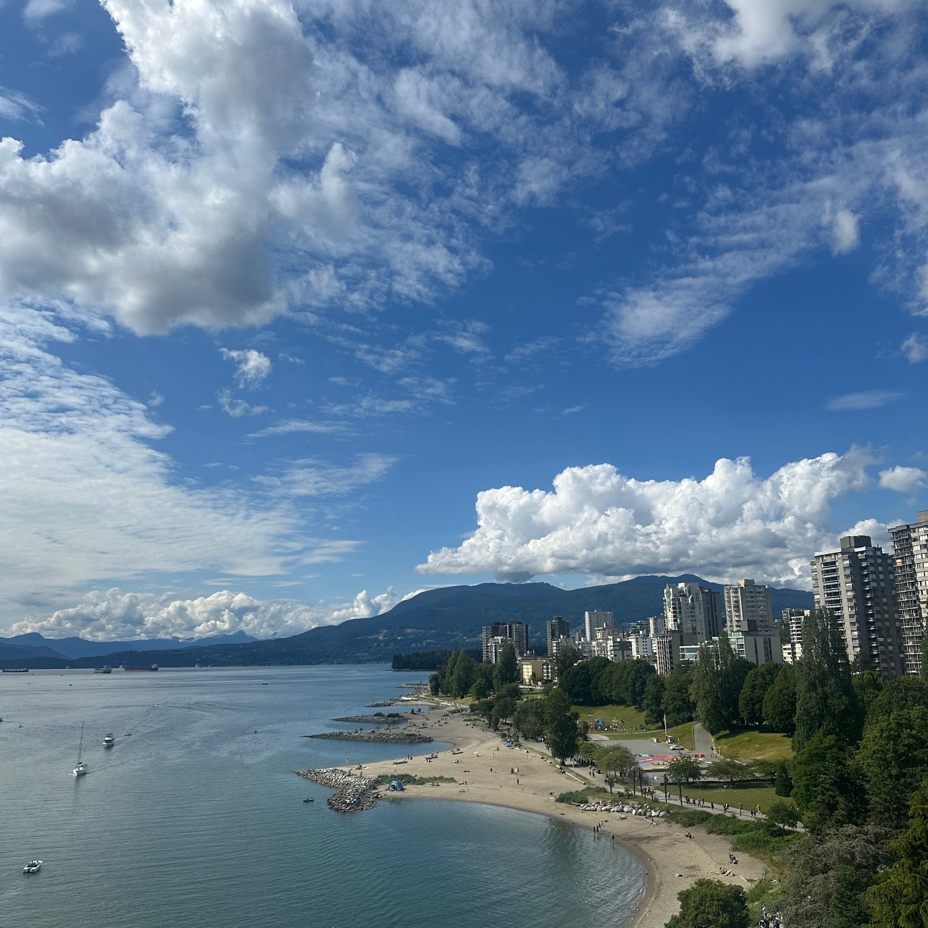 Vancouver cityscape with buildings by a beach and mountains under a blue sky with clouds.