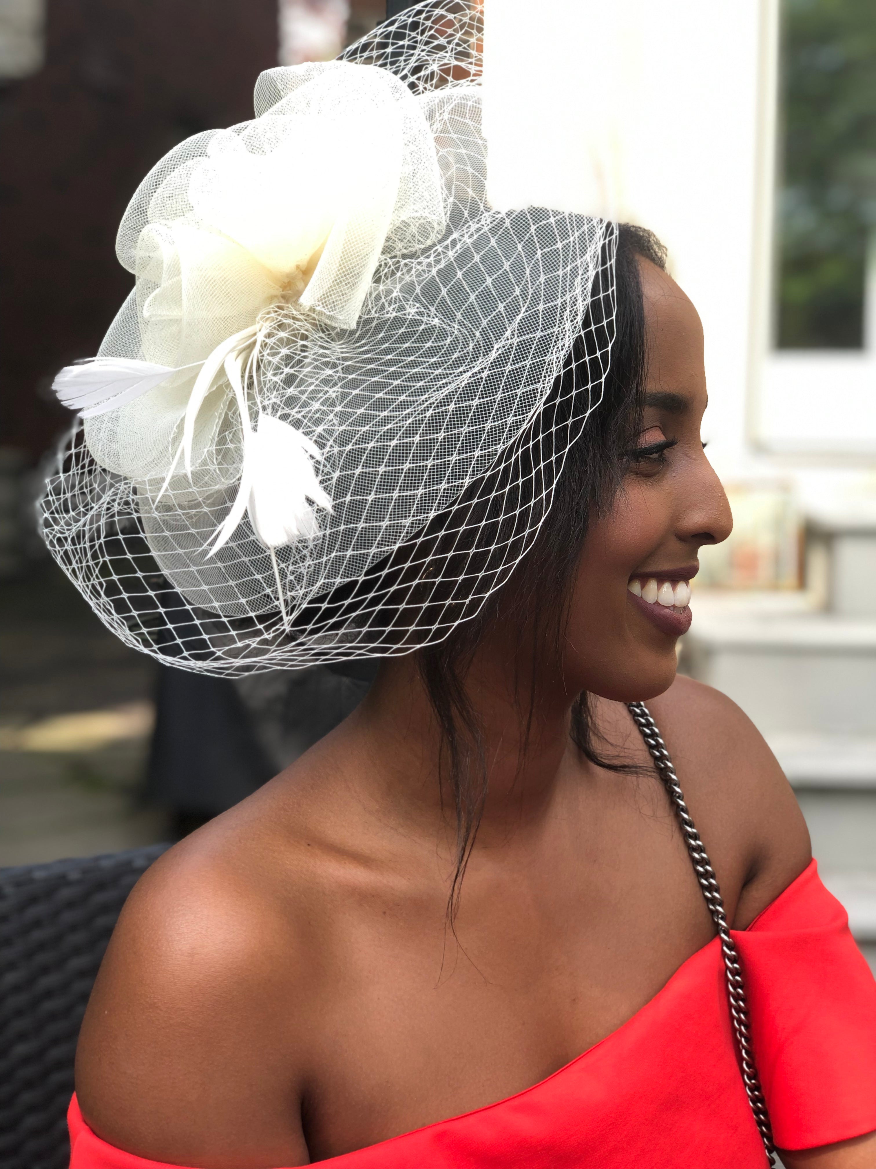 Woman wearing a decorative white hat with a veil and flowers, smiling outdoors.