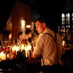 Barista working behind a counter with bottles and glasses in a dimly lit bar setting.