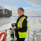 Person in a high-visibility vest standing on a boat with a large container ship in the background.