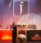 Woman in a black dress standing in front of a 'FOODBALL' branded backdrop with floral arrangements and drinks.