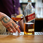 Person with tattoos holding a cocktail glass next to a bottle of Johnnie Walker Red Label whisky.