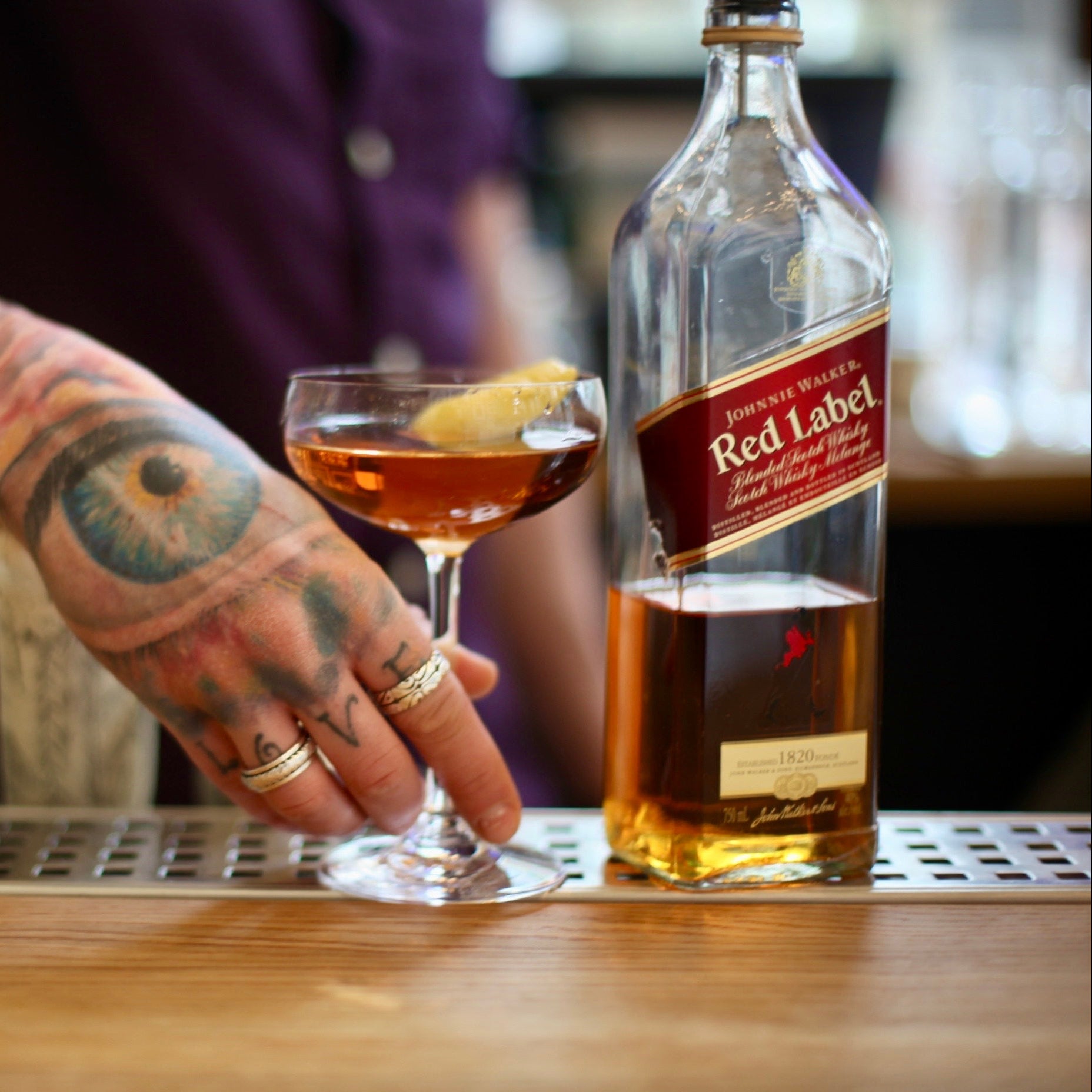 Person with tattoos holding a cocktail glass next to a bottle of Johnnie Walker Red Label whisky.